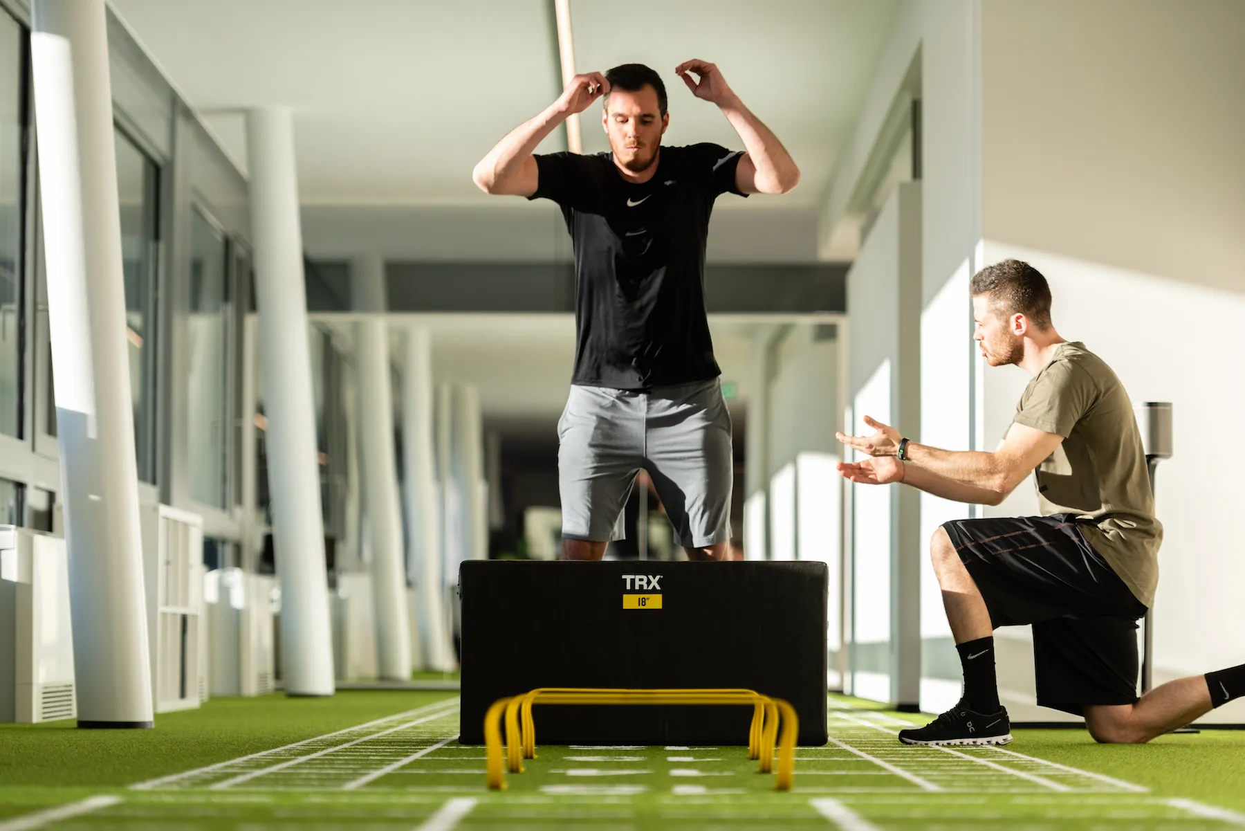 Sportler beim Sprungtraining über eine Box auf der Kunstrasenbahn, ein Trainer kniet daneben