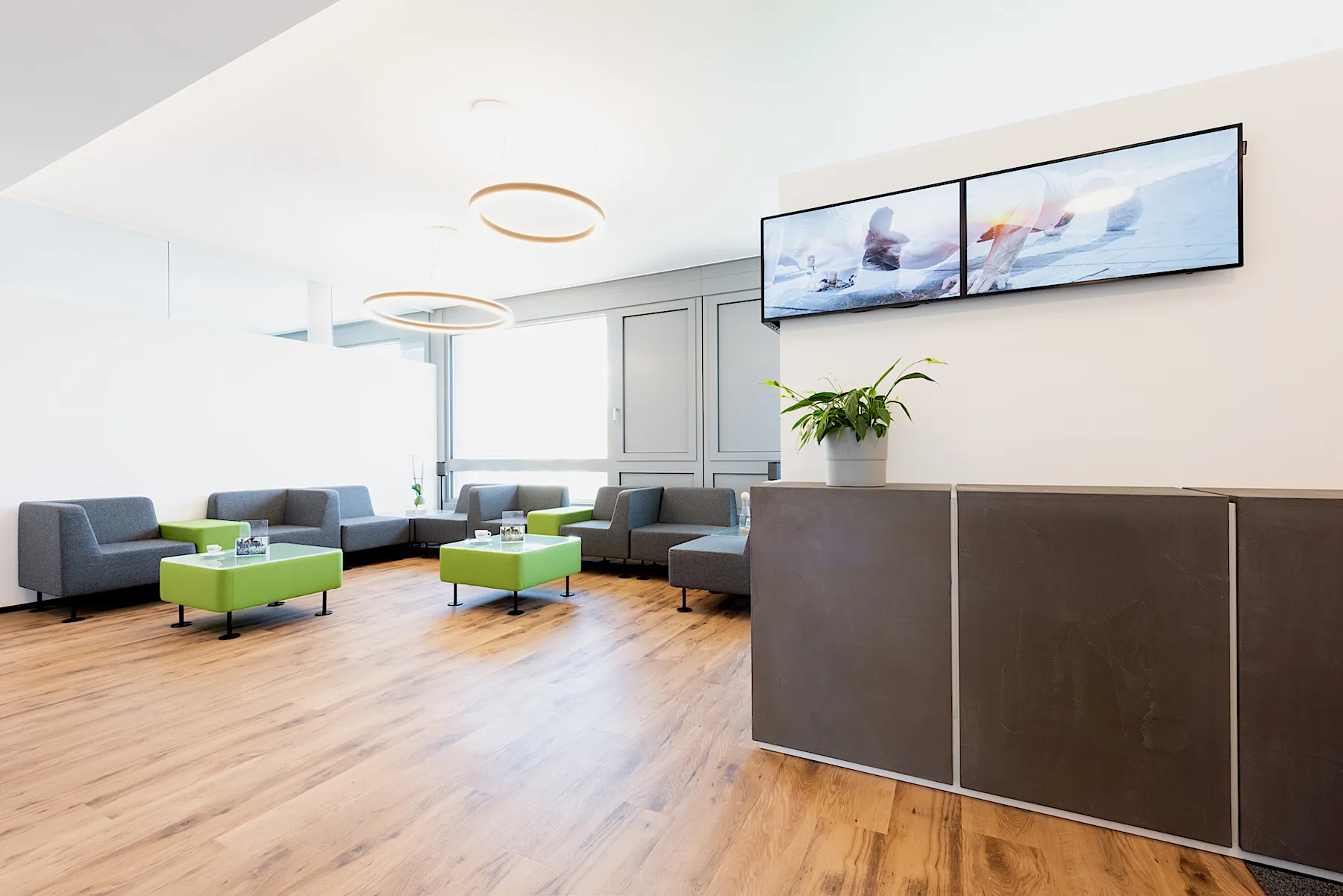 Wartebereich mit grauen Sofas, grünen Tischen und zwei großen Bildschirmen an der Wand